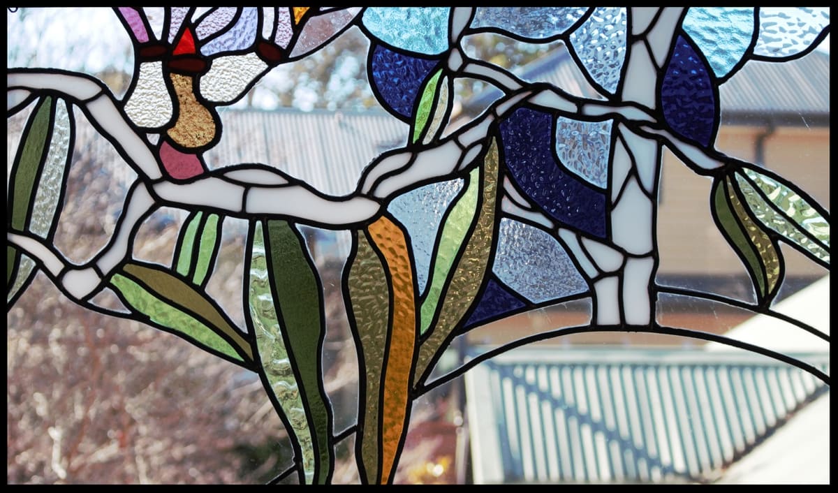 Angophora branch in leadlight with amber, plum and green leaves, a garden and roofline behind