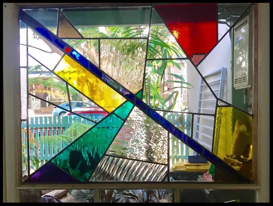 Modern leadlight window with a bold blue diagonal across fields of teal, purple and yellow, a garden behind